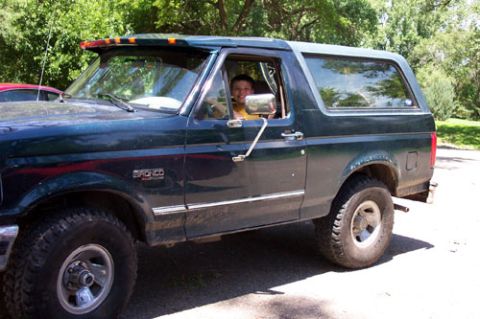 1995 Ford Bronco - Driver-side view of a dark green Ford Bronco with the removable hardtop in place, parked on a paved driveway with trees behind. It wears a…