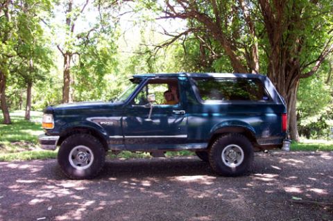 1995 Ford Bronco - A dark green two-door Ford Bronco full-size SUV with a removable hardtop, shown in profile from the driver's side, parked on a dirt road un…