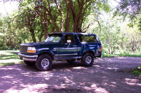 1995 Ford Bronco - Dark green 1990s full-size Ford Bronco parked on a shaded dirt road under leafy trees, seen from the front three-quarter angle. It sits on…