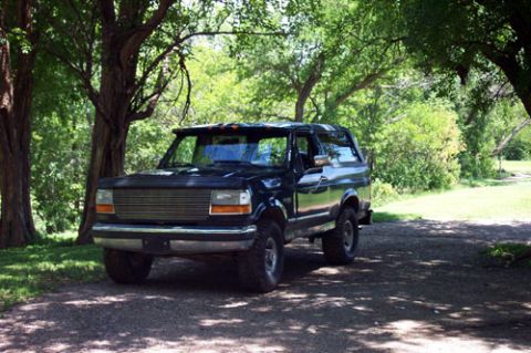 1995 Ford Bronco - A dark two-tone full-size Ford Bronco parked on a shaded gravel drive under large trees, viewed from the front three-quarter. It sits on a…