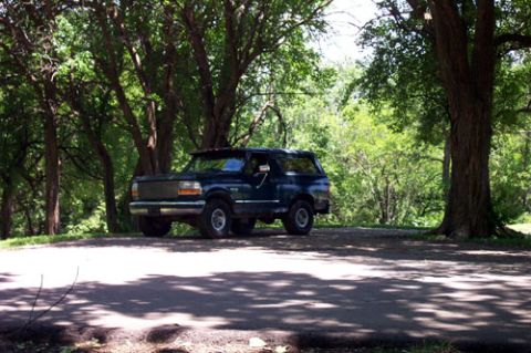 1995 Ford Bronco - A dark green two-door full-size Ford Bronco SUV parked at the edge of a shaded dirt clearing surrounded by large leafy trees. The truck is…
