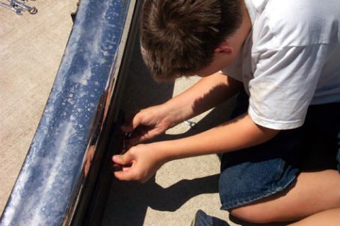 1995 Ford Bronco - A person in a white T-shirt and denim shorts kneels on a concrete driveway to work on a long dark metal bumper or step bar laid on the grou…