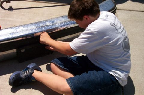 1995 Ford Bronco - A boy in a white T-shirt and denim shorts kneels on a driveway sanding a chrome truck bumper that has been removed and laid on its side, wi…