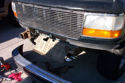 1995 Ford Bronco - Front end of a dark green 1990s Ford truck with the front bumper removed and lying face-down on the driveway, exposing the frame horns, tan…