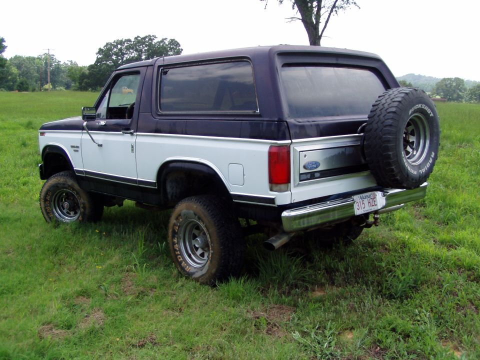 White and dark blue Ford Bronco with a spare tire mounted on the rear, parked in a grassy field