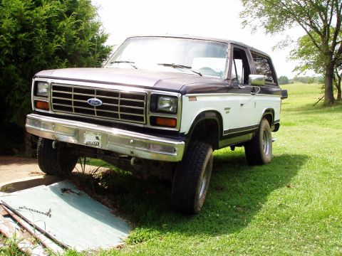1983 Ford Bronco - Three-quarter front view of a two-tone maroon-over-white full-size Ford Bronco with a removable hardtop, sitting in a grassy yard with its…