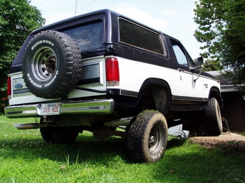 1983 Ford Bronco - Lifted 1980s Ford Bronco with a spare tire on the rear door, parked on grass.