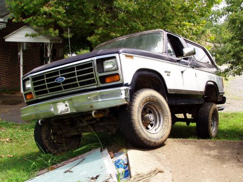1983 Ford Bronco - Low three-quarter front view of a lifted two-tone dark-over-white full-size Ford Bronco flexed with one front wheel dropped off a concrete…