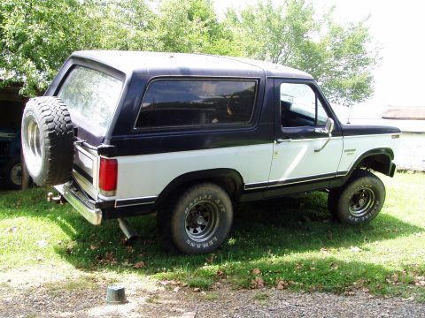 1983 Ford Bronco - Passenger-side three-quarter rear view of a lifted two-tone dark-over-white full-size Ford Bronco with hardtop, parked on grass beside a sh…
