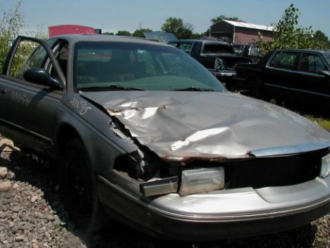 1997 Ford Taurus - A silver 1997 Ford Taurus sits in a junkyard with significant front-end damage and the driver's door ajar.