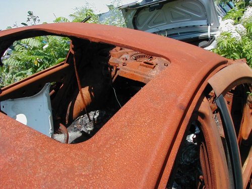Heavily rusted car interior with exposed dashboard and roof structure. Another car with open trunk visible in background