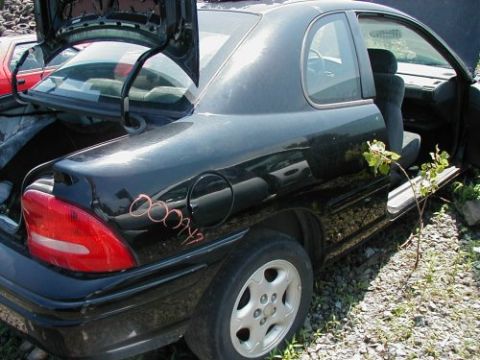 1997 Ford Taurus - A black sedan sits in a junkyard with its trunk and door open, showing signs of neglect and overgrowth.