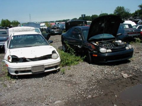 1997 Ford Taurus - Two damaged Dodge Neons sit in a junkyard under a clear sky.