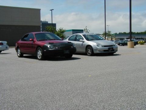 1997 Ford Taurus - Two cars, a red 1997 Ford Taurus and a silver Dodge Neon, are parked in a lot on a sunny day.