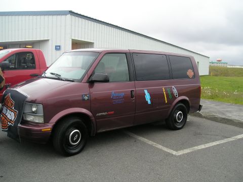 1996 Ford Bronco - A maroon van is parked next to a red truck, adorned with numerous brand stickers on its side.