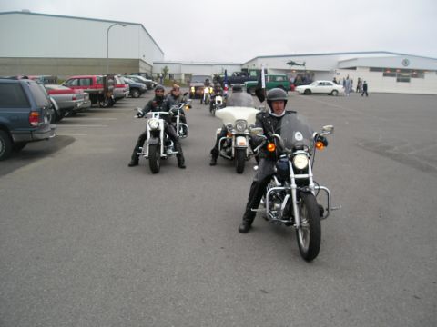 1996 Ford Bronco - A group of motorcyclists rides in formation across a paved area, with buildings and parked cars in the background.