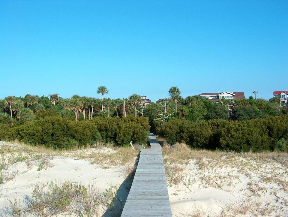 Wooden boardwalk leading through sand dunes and vegetation towards houses under a clear blue sky