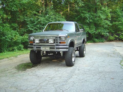 1986 Ford Bronco - A dark gray/green 1980s Ford Bronco full-size SUV shown from the front three-quarter angle, lifted on large off-road tires with chrome whee…