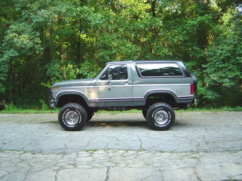 1986 Ford Bronco - A two-tone gray and silver 1980s Ford Bronco full-size SUV shown in side profile, lifted on chrome wheels with BFGoodrich All-Terrain T/A t…