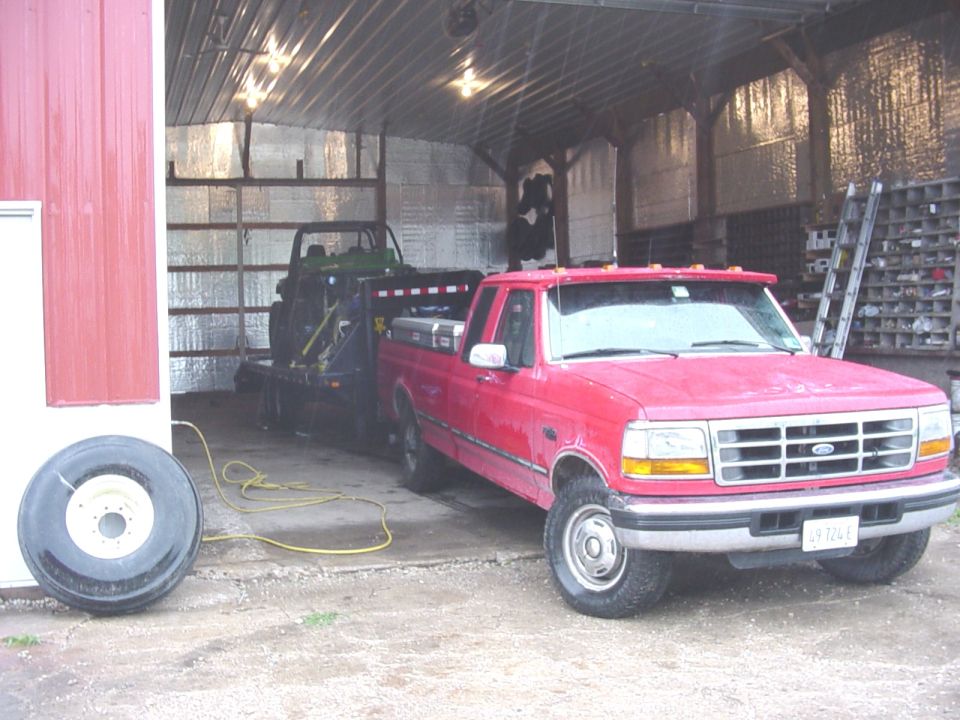 Red Ford F-Series truck, 1995 model year, towing a trailer with a green utility vehicle inside, parked in a barn