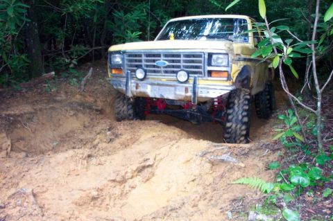 1985 Ford Bronco - Front three-quarter view of a lifted yellow Ford Bronco with a white top and red roll bar dropping into a deep clay rut on a narrow wooded…