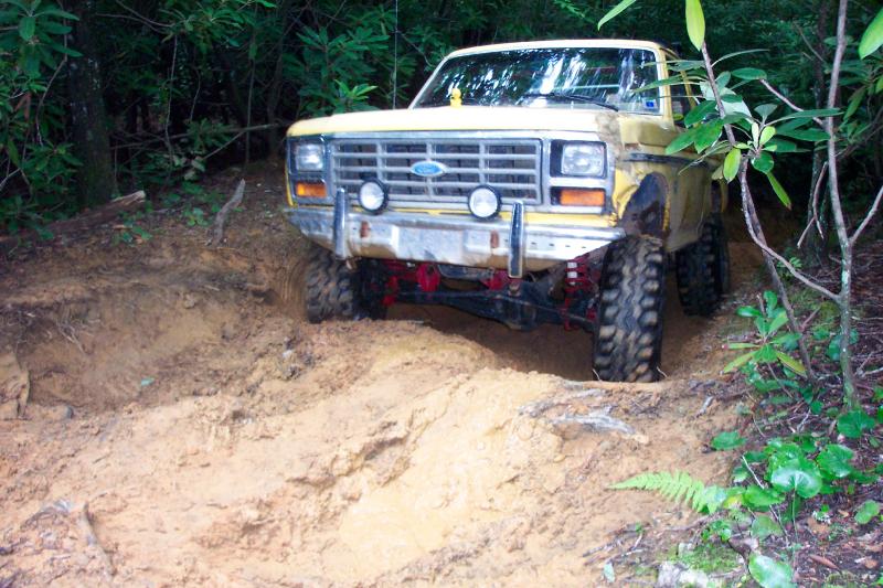 Lifted yellow Ford Bronco dropping into a deep clay rut on a narrow wooded trail
