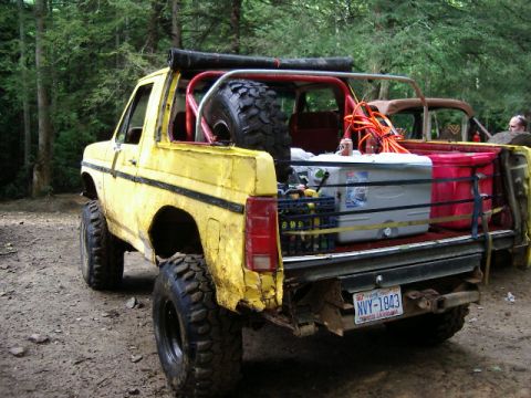 1985 Ford Bronco - Rear three-quarter view of a topless, lifted yellow Ford Bronco with a red roll bar, dented and scraped rear quarter panel, and a spare mud…