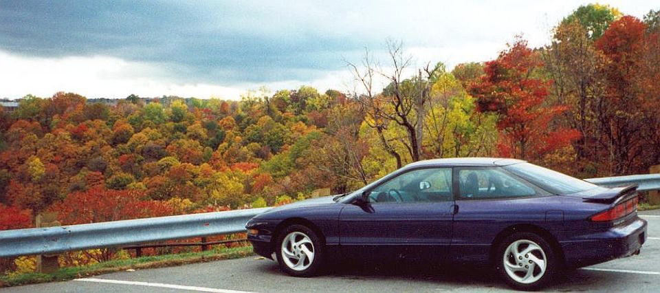 Purple-blue sport coupe with rear spoiler parked by a guardrail overlooking fall foliage