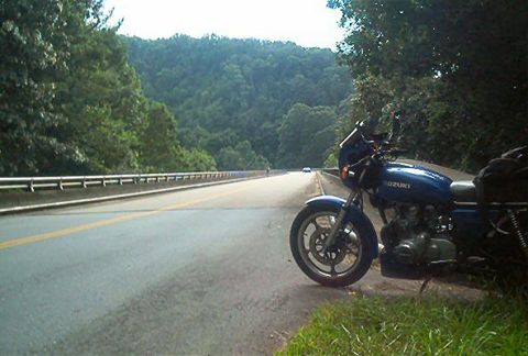 1985 Ford LTD Crown Victoria - A blue Suzuki motorcycle is parked on the side of a road with a wooded hillside in the background.