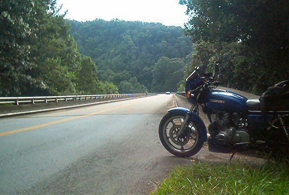 Blue Suzuki motorcycle parked on the shoulder of a highway with trees and hills in the background