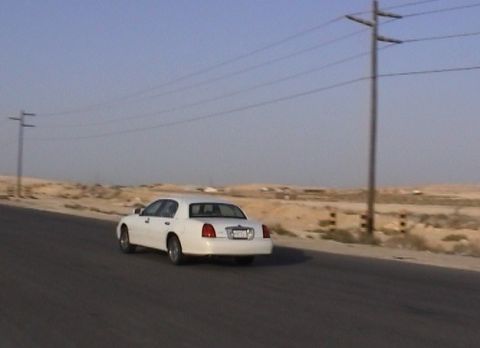 2002 Lincoln Town Car - A white 2002 Lincoln Town Car travels down a road in a dry, arid environment under a clear blue sky.