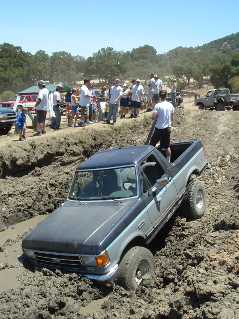 1989 Ford Bronco - A two-tone Ford Bronco navigates a muddy off-road course with spectators watching.