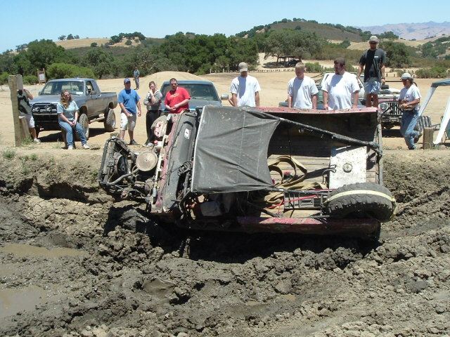 Maroon 4x4 with soft top rolled onto its side in a mud pit as spectators watch