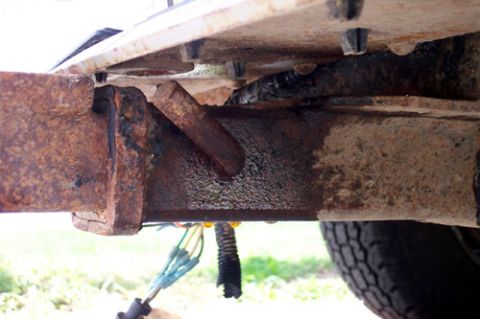 1995 Ford Bronco - Close-up under a truck's rear bumper showing heavily rusted bumper mounting brackets bolted to the frame rail, with flaking surface rust an…