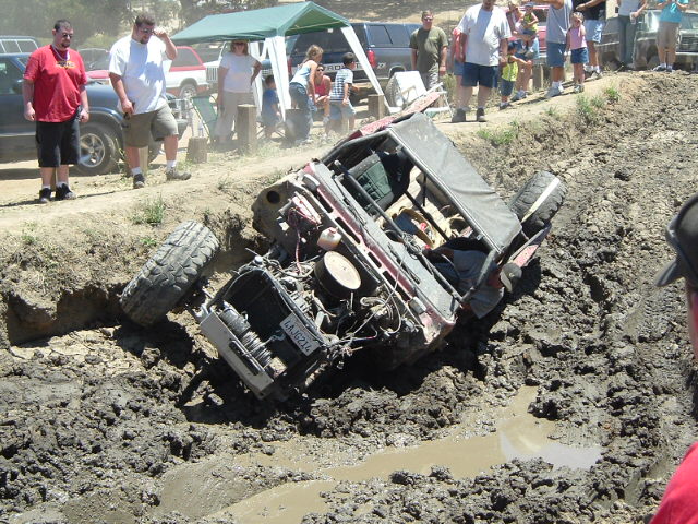 White SUV tipped on its side in a deep muddy off-road pit with spectators watching