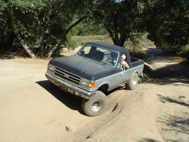 Topless two-tone Ford Bronco flexing up a dirt bank on a wooded trail, driver waving