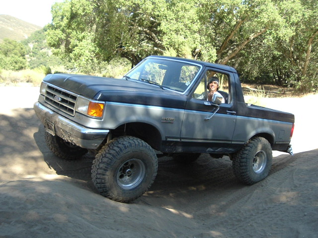 Lifted black-and-gray Ford Bronco on large tires climbing sandy dirt trail