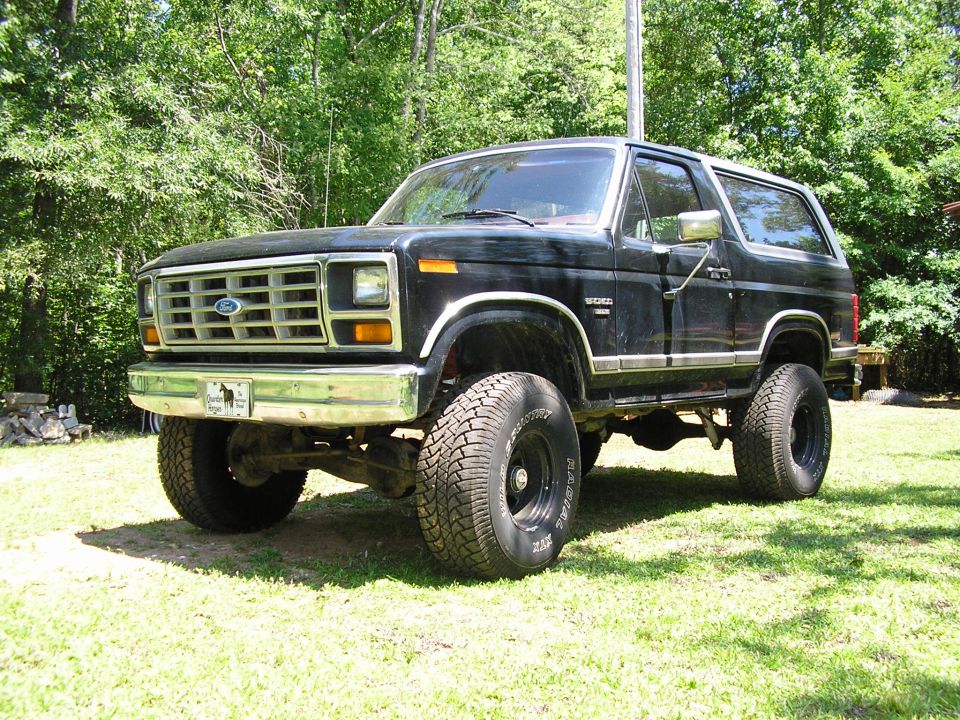 Black 1980s Ford Bronco SUV, lifted on big tires, parked on grass by trees.