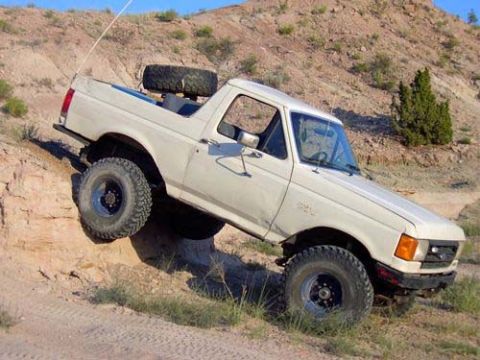 1987 Ford Bronco - A white 1987 Ford Bronco navigates a steep dirt incline, its off-road capabilities with large tires and a rear-mounted spare.