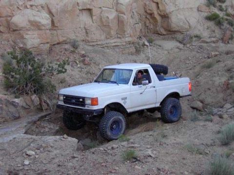 1987 Ford Bronco - A white Ford Bronco navigates a challenging off-road trail with steep, rocky terrain.