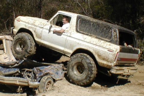 1978 Ford Bronco - A white 1978 Ford Bronco navigates a muddy obstacle course, crushing a smaller vehicle beneath its tires.
