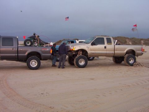 2000 Ford F-450 - Two lifted Ford trucks are parked on a sandy beach with a go-kart in the bed of one.