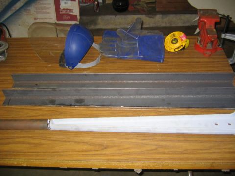 1978 Ford Bronco - Two lengths of metal C-channel or angle stock resting on a wood-grain workbench, alongside welding gear including a blue face shield, blue…