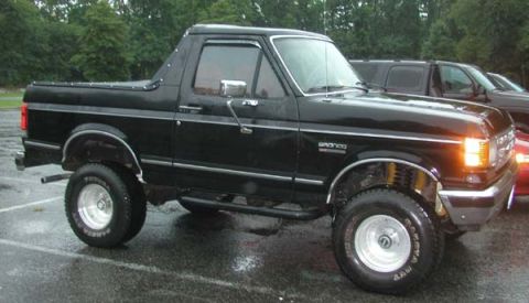 1988 Ford Bronco - A lifted black full-size Ford Bronco photographed from the driver's side front three-quarter angle on a wet parking lot, wearing a black so…