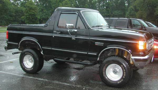 Lifted black Ford Bronco with black soft top on chrome wheels in a wet parking lot