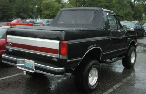 1988 Ford Bronco - Rear three-quarter view of a lifted black Ford Bronco on a wet parking lot, fitted with a black soft top over the cab and a snap-down tonne…
