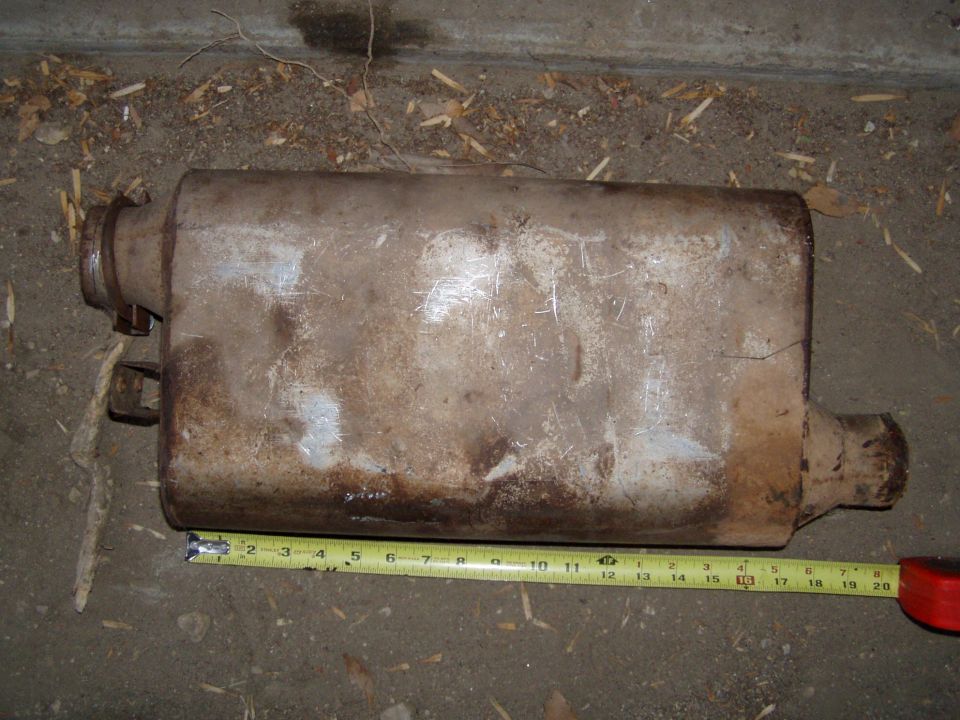 Rusty offset automotive muffler on concrete next to a yellow tape measure