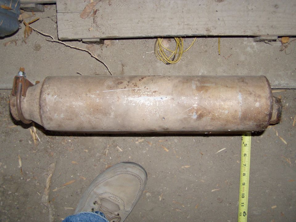 Rusty cylindrical muffler on concrete floor with tape measure and work boot for scale