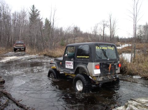 1980 Ford F-100 - Two off-road vehicles navigate a wet and muddy trail surrounded by trees.
