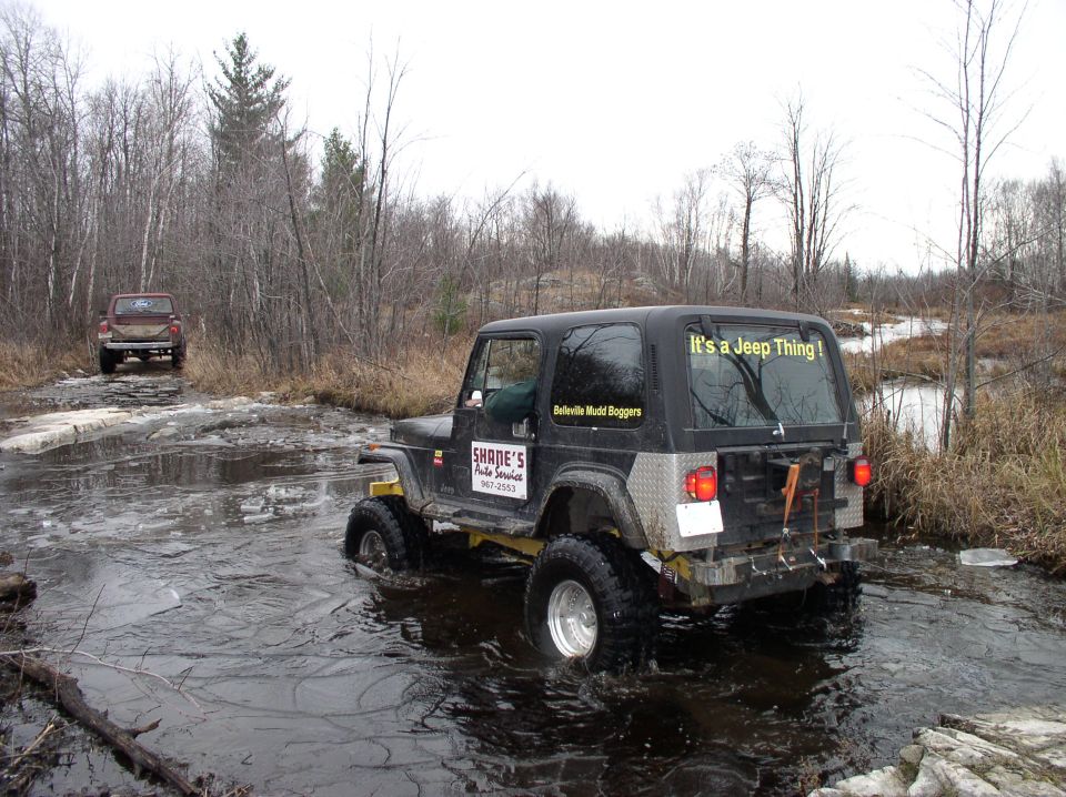 Jeep Wrangler driving through water with another truck behind it on a muddy trail